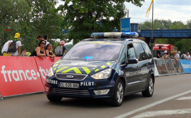 Polizeifahrzeug fährt an einer Menschenmenge vorbei, die Schilder hält, Bäume, eine Brücke, eine Flagge und einen bewölkten Himmel im Hintergrund.