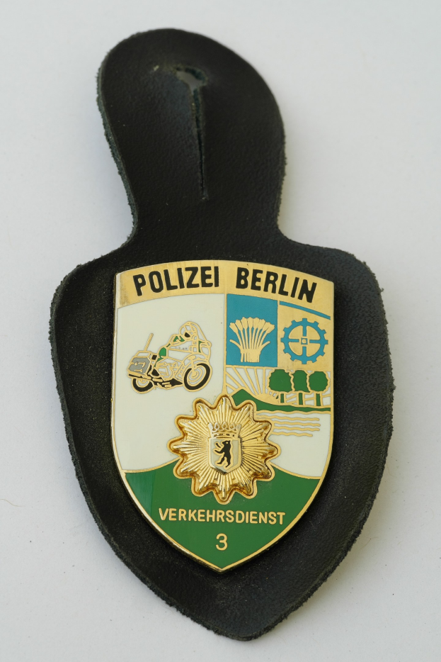 Ein schildförmiges Polizeibadgebnis mit der Aufschrift "Polizei Berlin" auf einem blauen Hintergrund, einem weißen Rand, einem weißen zentralen Stern und schwarzer fetter Schrift, das auf einer Oberfläche liegt.
