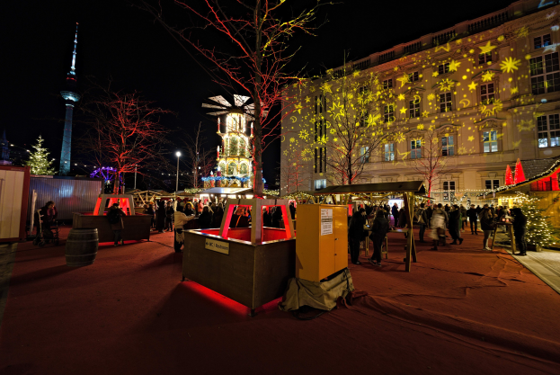 Ein belebter Weihnachtsmarkt in Berlin, Deutschland, mit Menschen um geschmückte Stände, festliche Lichter, Bäume, Gebäude, Laternenmasten und einen Turm im Hintergrund unter einem dunklen Himmel.