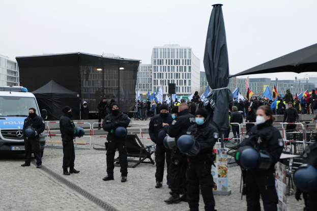Eine Gruppe von Polizisten steht vor einer Menge von Demonstranten in Berlin, Deutschland, mit Barrikaden, Zelten und Fahnen im Vordergrund und Gebäuden im Hintergrund unter einem klaren Himmel.