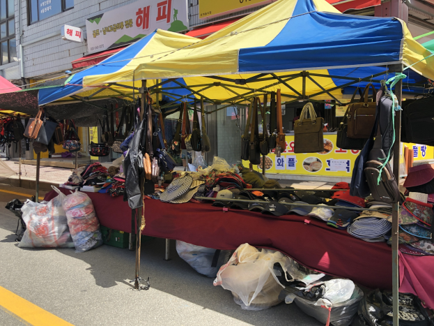 Ein Straßenmarkt mit Zelten, die Taschen, Hüte und andere Artikel präsentieren, vor Gebäuden mit Fenstern und Schildern im Hintergrund.