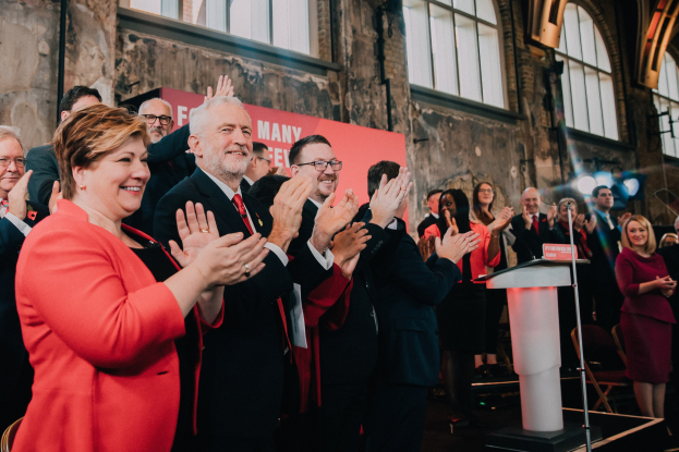 Eine Gruppe von Menschen, die vor einem Publikum klatschen, mit einem Podium, einem Mikrofon und einer Tafel mit Text auf der rechten Seite und Stühlen, einer Fahne, einer Wand, Fenstern und Lampen im Hintergrund.