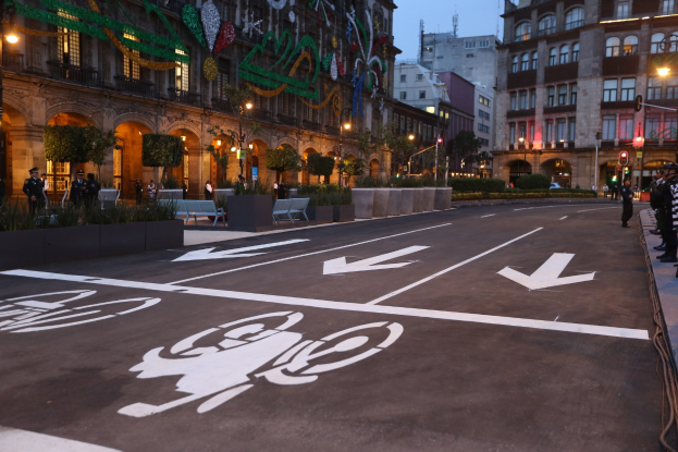 Stadtstraße mit einem ausgewiesenen Radweg, umgeben von Gebäuden, Straßenmöblierung, Fußgängern auf dem Gehweg, Grünflächen und einem klaren blauen Himmel.