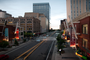 Eine Stadtlandschaft mit einer Straße voller Autos unten, flankiert von Gebäuden und Wolkenkratzern auf beiden Seiten, unter einem Himmel oben.