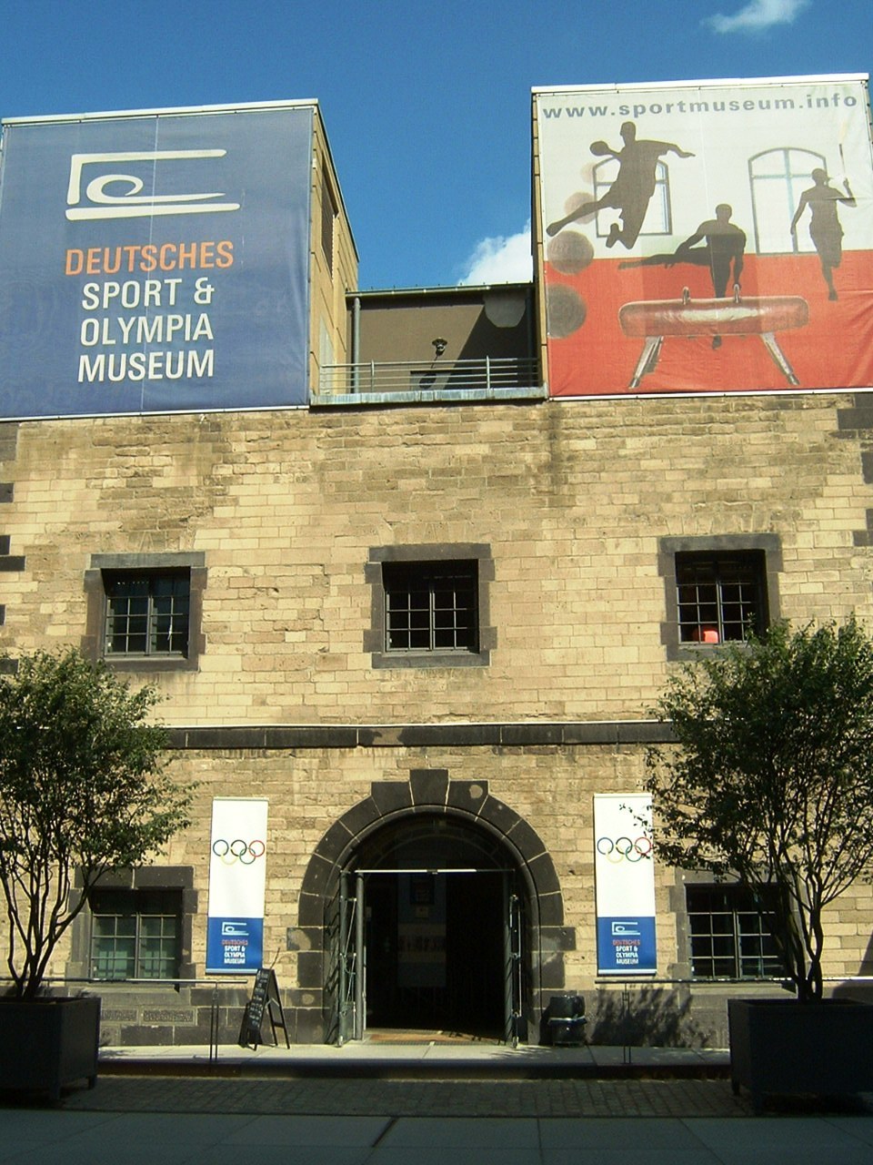 Das Deutsche Sport & Olympische Museum in Berlin, Deutschland, ein Gebäude mit Fenstern und einer Tür, umgeben von Bäumen und Bannern mit Text und Bildern, unter einem bewölkten Himmel.