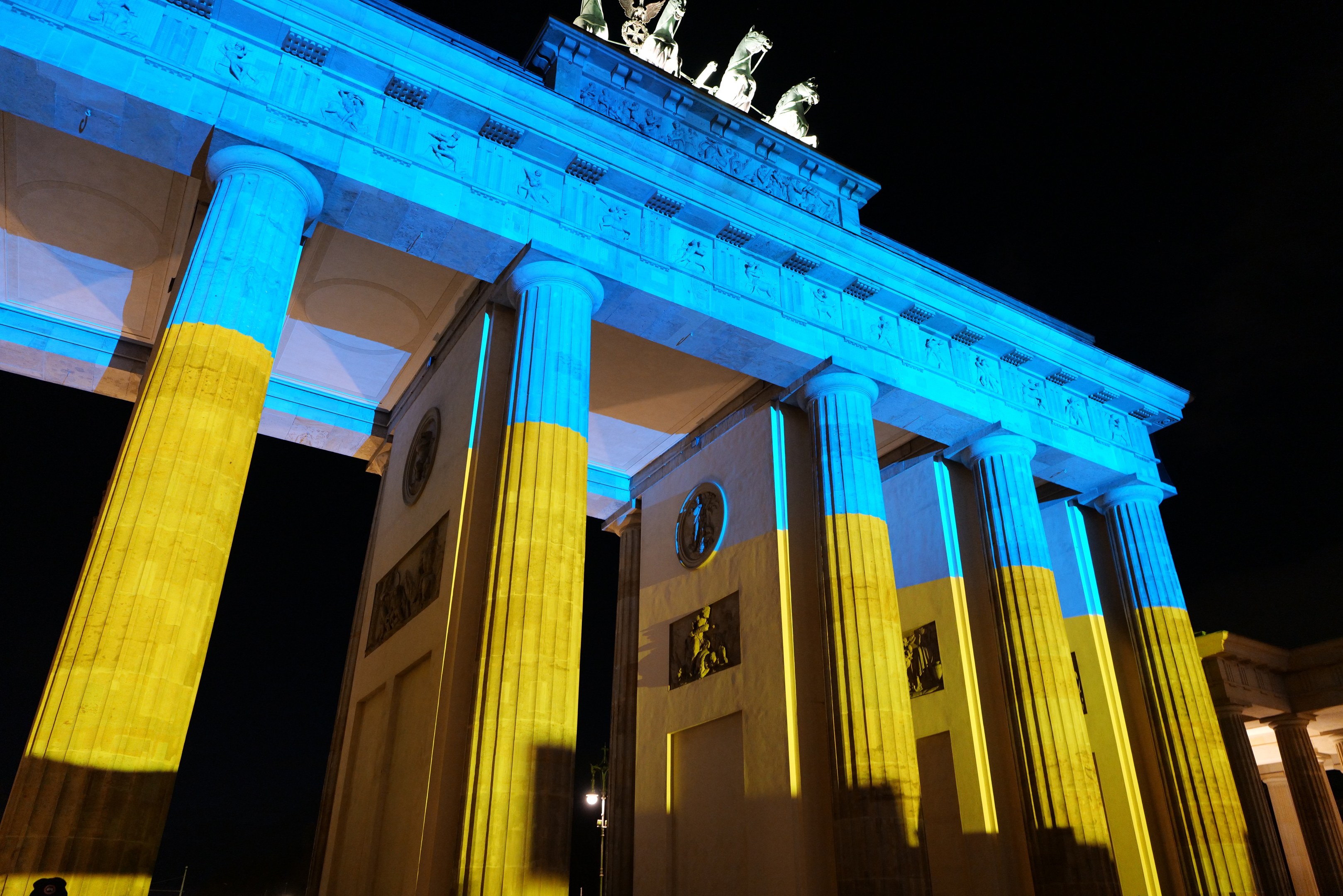 Brandenburger Tor in Berlin in Blau und Gelb beleuchtet, mit Skulpturen gegen einen dunklen Hintergrund