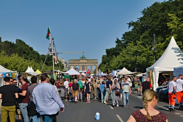 Eine Menschenmenge geht eine Straße mit Zelten, Fahrzeugen und Bäumen entlang, mit einem Bogen und einem klaren blauen Himmel im Hintergrund und Fahnenmasten auf der linken Seite, die wahrscheinlich ein Oktoberfest-Event in München, Deutschland, kennzeichnen.