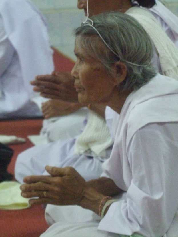Alte Frau liegt im Krankenhausbett mit gefalteten Händen, geschlossenen Augen und Kopf auf den Händen ruhend, umgeben von Menschen im Hintergrund.