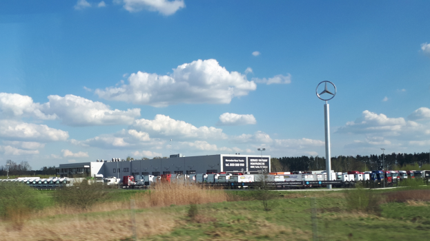 Ein Mercedes-Benz-Werk in Deutschland mit Fahrzeugen auf der Straße, umgeben von Gras, Pflanzen, Bäumen, Gebäuden und Polen unter einem bewölkten Himmel.