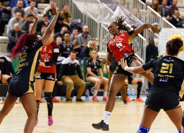 Eine Gruppe von Frauen, die Volleyball auf einem Feld mit einem Ball in der Mitte spielt, während Zuschauer auf Stühlen und Geländern im Hintergrund sitzen und Flaschen auf dem Boden liegen.