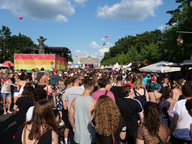 Eine gro├če Menge geht eine Stra├če entlang, die mit Zelten, B├Ąumen, Pfosten, Lichtern und einer Statue ges├Ąumt ist, w├Ąhrend des Berliner Pride-Festivals, mit Geb├Ąuden und einem Himmel voller Wolken und Ballons im Hintergrund.