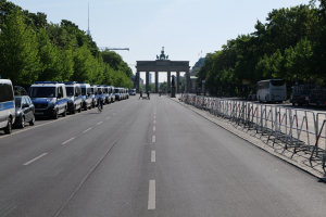 Lange Reihe von Polizeiwagen am Straßenrand vor dem Brandenburger Tor mit Menschen auf Fahrrädern und auf der Straße, Barrieren, Bäume, ein Bogen mit Statuen im Hintergrund und sichtbarer Himmel.