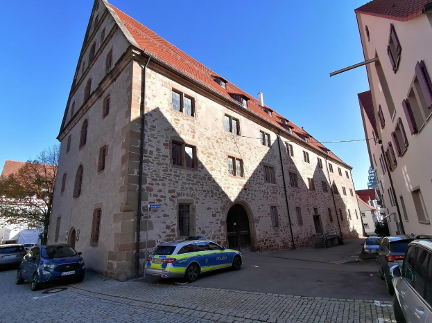 Polizeiauto vor einem Gebäude mit Fenstern geparkt, umgeben von Bäumen und einem klaren blauen Himmel.