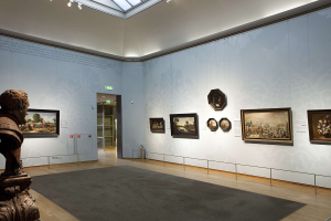 Ein großer Raum mit einer Statue links, einem Teppichboden, gerahmten Kunstwerken an den Wänden, einer Glasdecke mit Einbaulichtern und Türen im Hintergrund, Teil des Kunsthistorischen Museums in München, Deutschland.