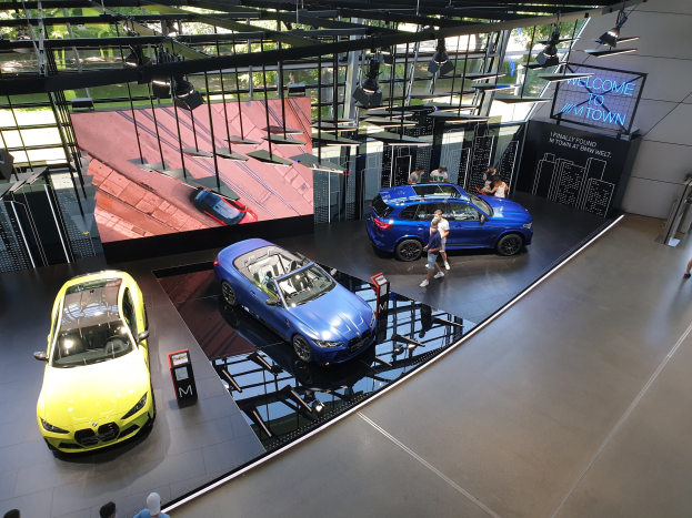 Eine Gruppe von Menschen steht im BMW Museum in München, mit Autos auf der linken Seite und einem Schild auf der rechten Seite, das 'BMW Museum München' lautet, mit Blick auf Bäume, Gebäude und den Himmel durch Glasfenster.