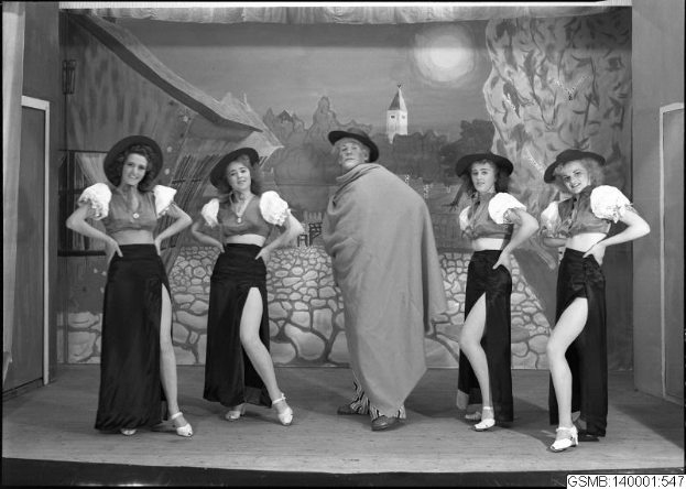 Fünf Frauen mit Hüten stehen auf einer Bühne, lächeln, mit einem Gemälde und einer Tür im Hintergrund, schwarz-weiß, 1940er Jahre.