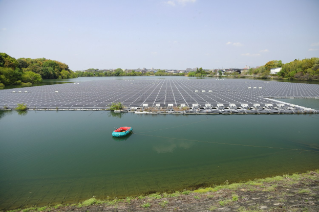 Kleines Boot mit Solarpanelen auf einem Gewässer treibend, umgeben von Grünflächen, mit Gebäuden und einem klaren blauen Himmel im Hintergrund.