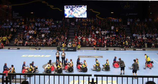 Eine Gruppe von Menschen in Rollstühlen auf einem Feld bei den Invictus Games Düsseldorf 2020, umgeben von einer Menge, mit Stufen zu einem Bildschirm und Texttafeln im Hintergrund.