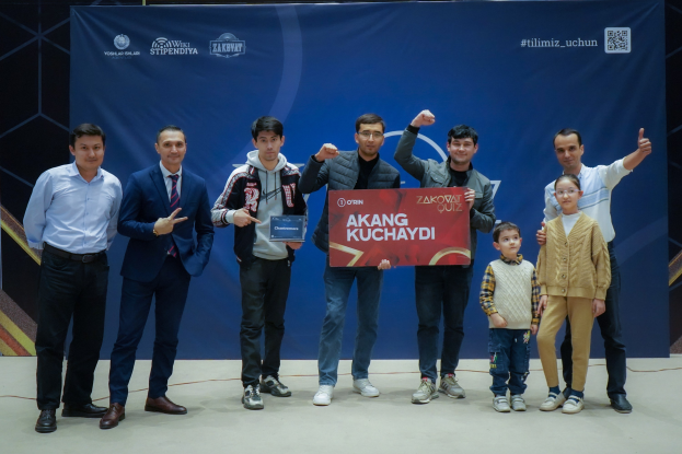 Gruppe von Menschen auf einer Bühne mit einer Tafel, auf der "Akang Kuchaydi - Zakovat Quiz" steht, mit einem Banner, Lichtern und anderen Gegenst├Ąnden im Hintergrund.