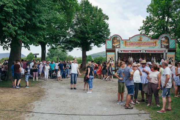 Menschenmenge an einem Oktoberfest-Essenstand in München, Deutschland, mit einigen, die Mützen und Okulare tragen, andere halten Gegenstände, auf Gras mit Bäumen, Hügeln und einem bewölkten Himmel im Hintergrund.