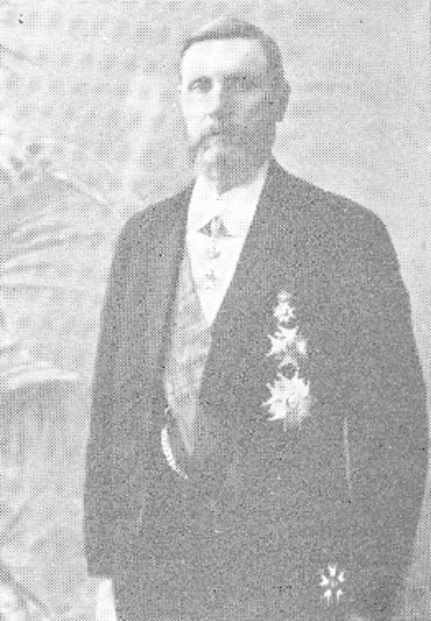 Ein Mann in einem schwarzen und weißen Anzug mit einem weißen Hemd und schwarzer Krawatte steht vor einer üppigen grünen Pflanze und sieht ernst aus.