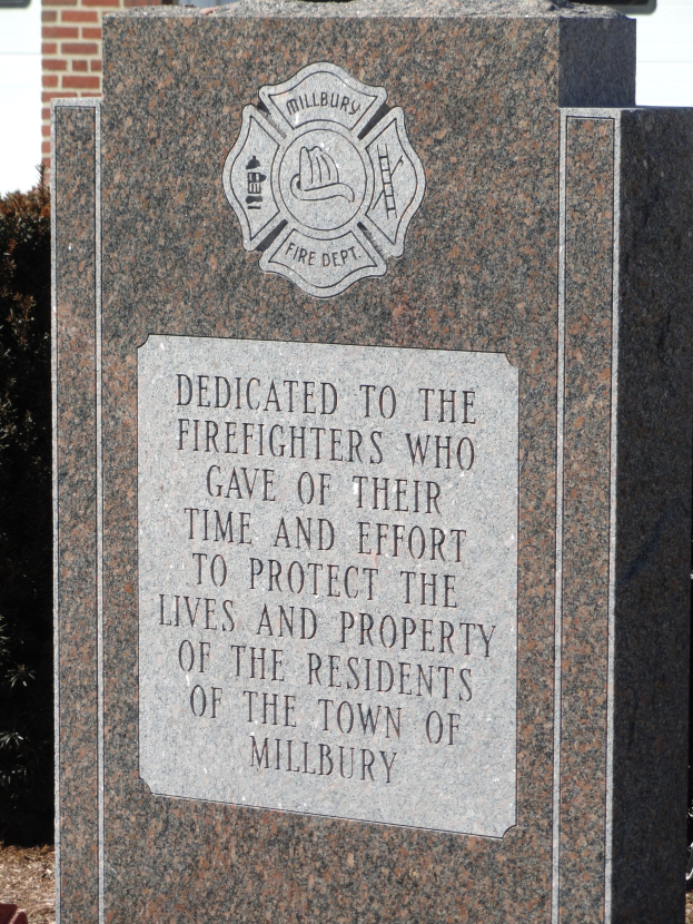 Gedenkstein mit einer Plakette, gewidmet an Feuerwehrleute in Millbury, aufgestellt vor einem Backsteingebäude mit umliegenden Pflanzen und Bäumen.