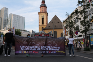 Eine Gruppe von Menschen in Masken, die eine Straße entlanggehen und ein Transparent halten, mit einem geparkten Auto auf der linken Seite, Gebäuden und Bäumen im Hintergrund und einem klaren blauen Himmel.