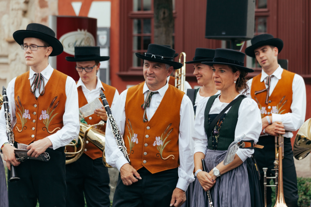 Eine Gruppe von Menschen in traditioneller bayerischer Tracht, die Musikinstrumente halten, vor einem Gebäude mit Fenstern, einem Lautsprecher und Pflanzen im Hintergrund.