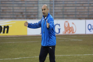 Ein Mann in einer blauen Jacke steht auf einem Fußballfeld, umgeben von Tafeln mit Text und Logos, und sieht konzentriert während eines Trainings.