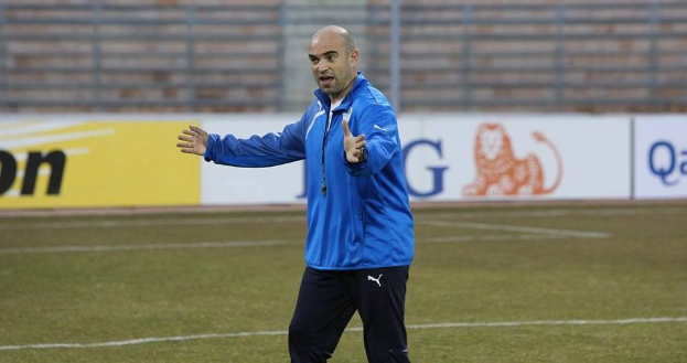 Ein Mann in einer blauen Jacke steht auf einem Fußballfeld, umgeben von Tafeln mit Text und Logos, und sieht konzentriert während eines Trainings.