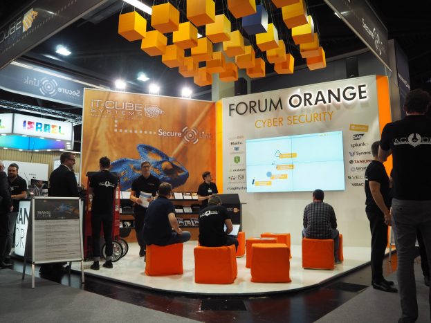 Eine Gruppe von Menschen an einem Messestand für IT-Cube Systems Forum Orange Cyber Security, mit einigen sitzend und anderen stehend, um eine Anzeige mit Text, einen Bildschirm, Regalen und Schildern unter Deckenbeleuchtung.