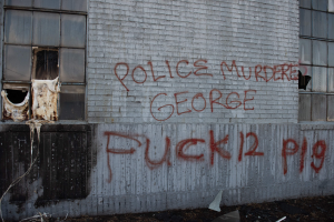 Graffiti an einer Gebäudewand mit der Aufschrift "Polizeimord George Fuck 12 P19" mit Fenstern und verstreuten Gegenständen davor.