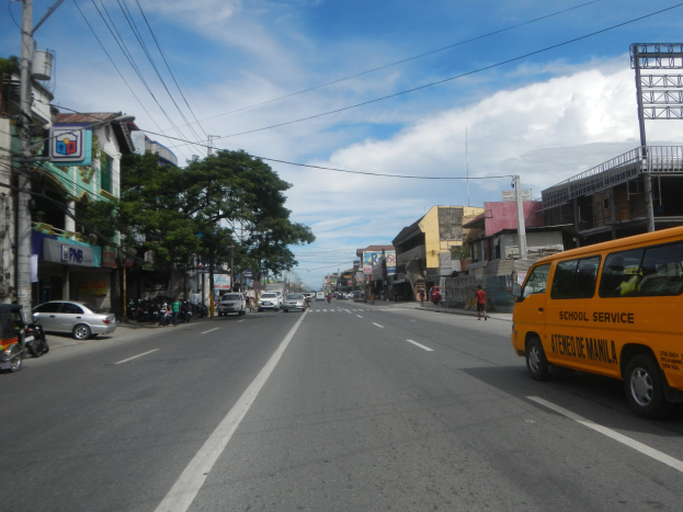 Ein gelber Schulbus fährt eine städtische Straße mit hohen Gebäuden, Fußgängern auf den Bürgersteigen, Bäumen, Strommasten mit Drähten und Texttafeln entlang, unter einem bewölkten Himmel.