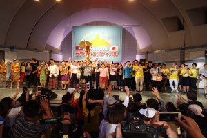 Gruppe von Menschen auf einer Bühne mit Kameras, Handys und Musikinstrumenten während einer Festivaleröffnung mit einem Banner, Wänden und Deckenlampen im Hintergrund.