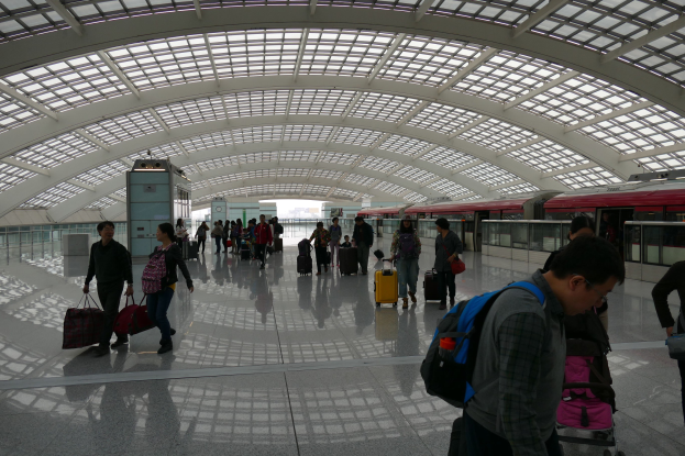 Menschen mit Gepäck durch den Flughafen gehen, einige tragen Taschen und Koffer, ein Zug ist auf der rechten Seite zu sehen und eine Glaswand im Hintergrund.