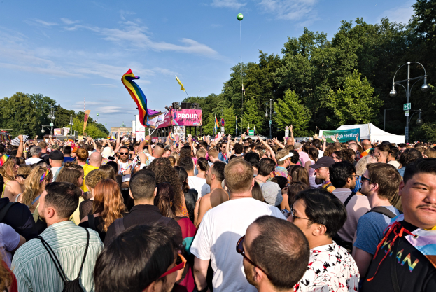 Große Menschenmenge vor einem Gebäude, viele halten Regenbogenflaggen und Banner, mit einem Zelt, Laternenmasten, Bäumen und einem schwebenden Ballon im Hintergrund.