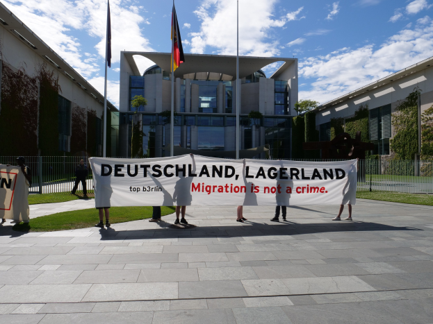 Gruppe von Menschen mit einer Fahne, auf der 'Deutschland, Lagerland Migration ist kein Verbrechen' steht, vor einem Gebäude mit einem Metallzaun, umgeben von Gras, Pflanzen, Bäumen und Fahnen.