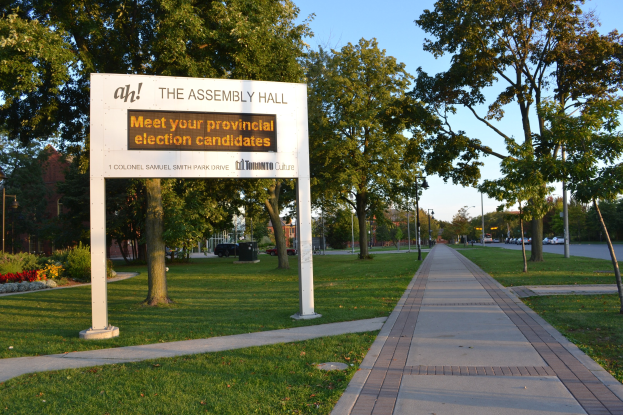Board auf Gras mit Text "Die Versammlungshalle - Treffen Sie Ihre Landtagswahlkandidaten" umgeben von Weg, Bäumen, Blumen, Straßenlaternen, Fahrzeugen, einem Gebäude und einem bewölkten Himmel.