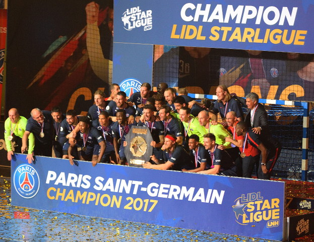 Spieler von Paris Saint-Germain feiern mit dem Ligue-1-Pokal, tragen Medaillen, vor einer Tafel mit Text und Logos, mit einem Netz und einem Bildschirm im Hintergrund.