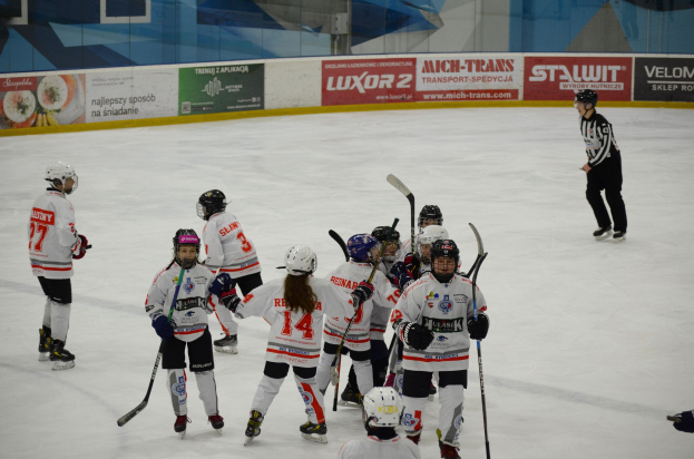 Eine Gruppe junger Menschen, die auf einem Eisplatz Eishockey spielt und Helme trägt sowie Hockey-Schläger in den Händen hält, mit einer Person auf der rechten Seite und Texttafeln im Hintergrund.