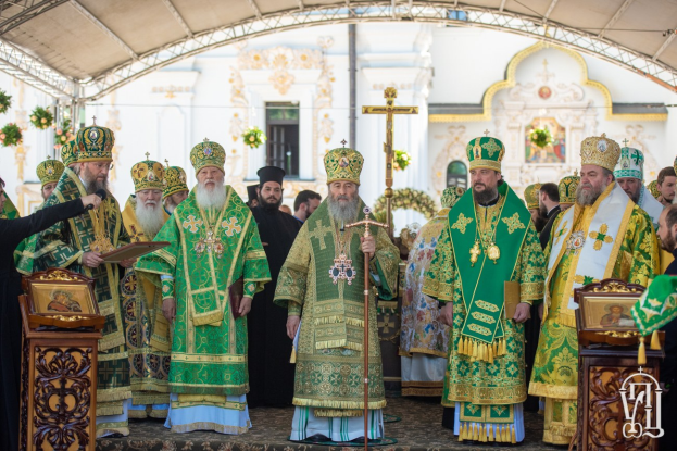 Gruppe von Priestern vor einer Kirche während einer religiösen Zeremonie, wobei einer ein Buch und ein Mikrofon hält, Kreuzsymbol und Blumen im Hintergrund sichtbar