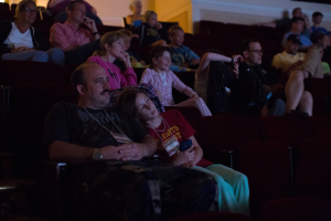 Eine Gruppe von Menschen sitzt in einem Kino mit einem Bildschirm auf der rechten Seite und einer Wand im Hintergrund.