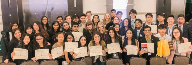 Gruppe von Menschen mit Urkunden in einem Raum mit Stühlen und einer Wand im Hintergrund, wahrscheinlich feiern sie eine Abschlusszeremonie für das akademische Jahr 2019-2020.