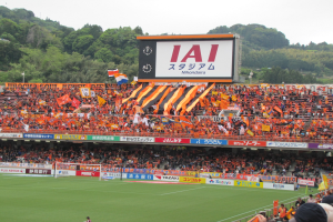 Ein Fußballspiel auf einem grünen Feld mit einem Tor, umgeben von einer großen Zuschauermenge in den Stadionrängen, Bannern, Fahnen, einem großen Bildschirm, Bäumen und einem klaren blauen Himmel.