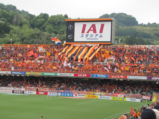 Ein Fußballspiel auf einem grünen Feld mit einem Tor, umgeben von einer großen Zuschauermenge in den Stadionrängen, Bannern, Fahnen, einem großen Bildschirm, Bäumen und einem klaren blauen Himmel.