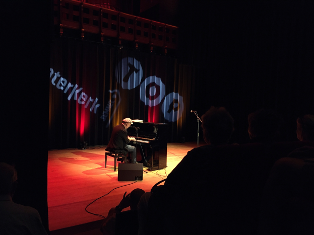 Ein Mann mit Mütze sitzt an einem Piano auf einer schwach beleuchteten Bühne, mit einem Mikrofon auf der rechten Seite und einem Vorhang mit Text im Hintergrund.