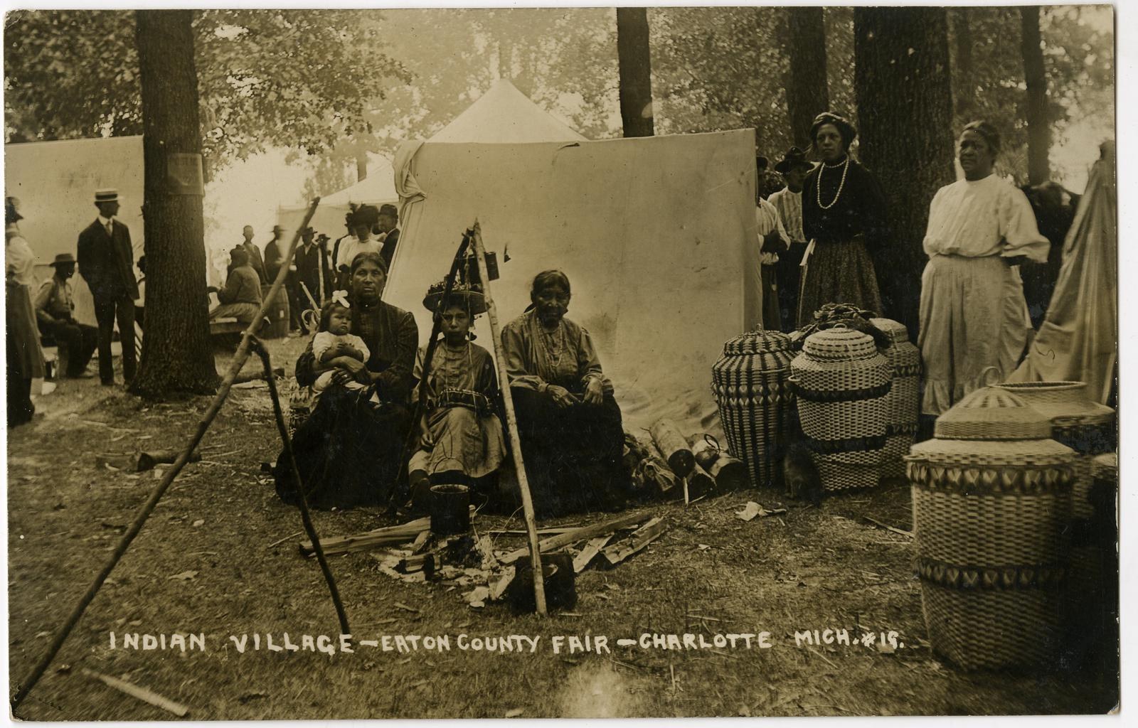Schwarz-weiß-Foto einer Gruppe von Menschen, die sich um ein Zelt auf dem Eton County Fair in Charlotte, North Carolina, versammeln, mit Körben und Gegenständen rechts, Bäumen und einer Wand im Hintergrund und Text unten.