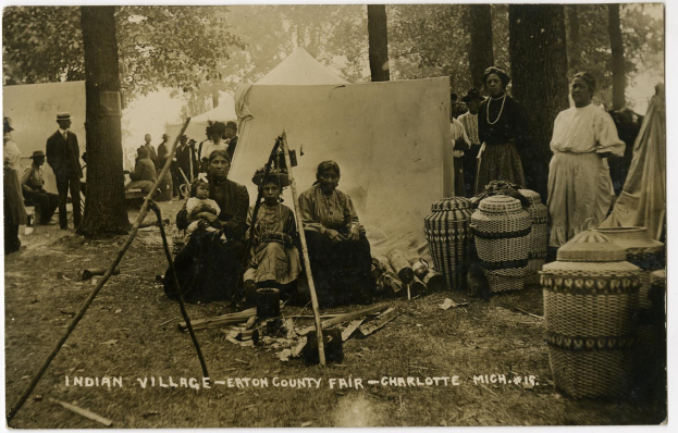 Schwarz-weiß-Foto einer Gruppe von Menschen, die sich um ein Zelt auf dem Eton County Fair in Charlotte, North Carolina, versammeln, mit Körben und Gegenständen rechts, Bäumen und einer Wand im Hintergrund und Text unten.