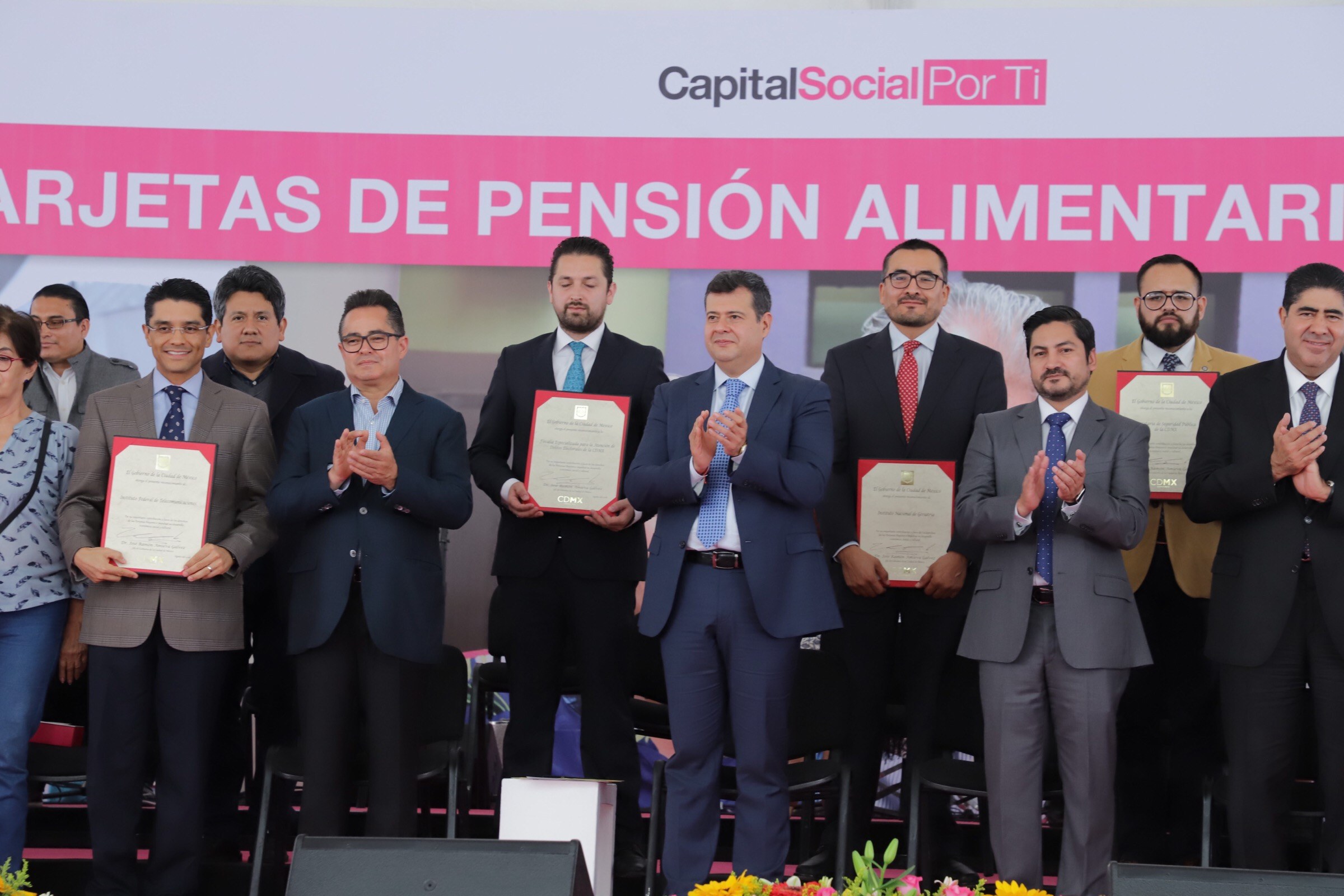 Gruppe von Menschen auf einer Bühne, die klatschen und Zertifikate halten, mit Stühlen in einem Halbkreis, einem Banner mit der Aufschrift "Capital Social for Ti" im Hintergrund und Blumenbouquets und Lautsprechern unten.