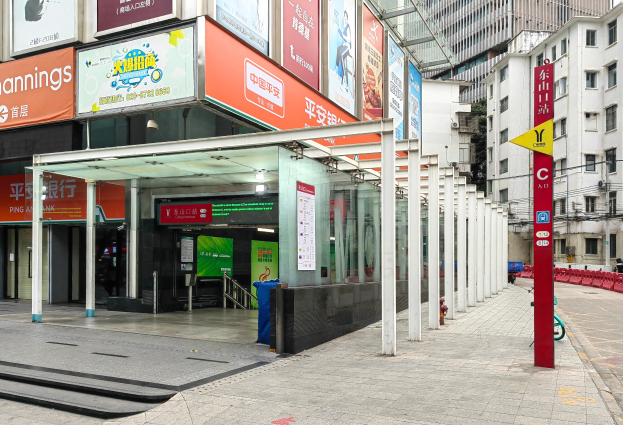 Eine Stadtstraße mit einer Bushaltestelle, Gebäude auf beiden Seiten, eine Straße mit einem Müllcontainer, rote und gelbe Schilder mit Text, Menschen auf dem Gehweg und eine Rolltreppe auf der linken Seite.